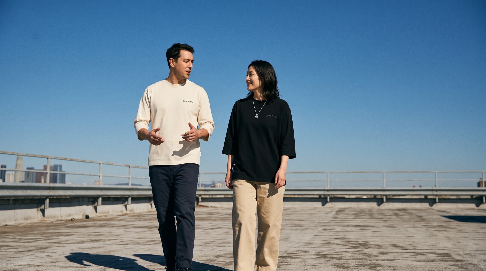 Two people walking on a rooftop under a blue sky, wearing good-tone tees